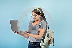 Cheery Indian teen girl with backpack and laptop pc having online lesson on blue studio background
