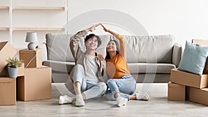 Cheery millennial Asian couple relocating to new home, making house roof gesture, sitting on floor among carton boxes