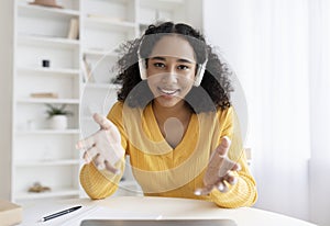 Cheery young black female wearing headphones, using laptop, working or studying online, having business meeting at home