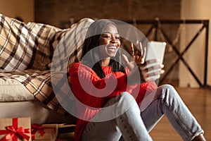 Cheery young black lady in winter outfit waving at smartphone camera, having video chat with friend at home