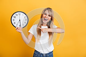 Cheery young blonde woman posing isolated over yellow wall background dressed in white casual t-shirt holding clock pointing