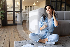 Cheery young Caucasian lady in earphones listening to music or audio book on smartphone, sitting on floor at home