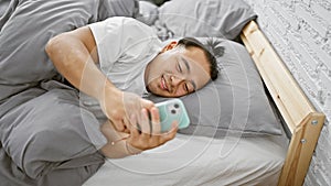 Cheery young chinese man in pyjamas luxuriating in a cozy bedroom, using smartphone for fun morning texting, confident asian