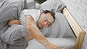 Cheery young chinese man in pyjamas luxuriating in a cozy bedroom, using smartphone for fun morning texting, confident asian adult