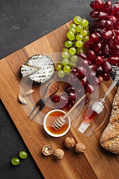 Cheese head, bunch of grapes, honey, nuts and wineglass on wooden board and black background