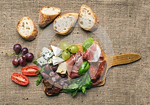 Cheese and meat platter with fresh grapes, cherry-tomatoes, olives, herbs and bread pieces on a rustic wood board over the rough