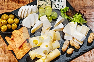 Sýrový talíř, sada různých sýrů na černém talíři s hrozny a zelenými olivami hranolky a ořechy