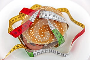 A cheeseburger sitting on a plate with multi-colored measuring tape twining around it. A subtle reminder to stick to a healthy