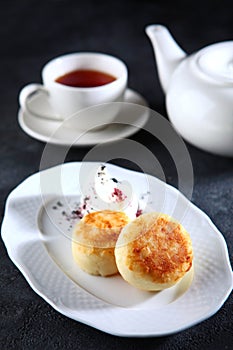 Cheesecakes on a white plate. Tea in a white porcelain teapot. Dark background. The concept of breakfast.