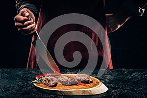 Chef in apron with meat fork and knife slicing gourmet grilled steaks with rosemary and chili pepper on wooden board