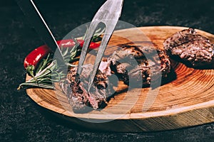 Chef in apron with meat fork and knife slicing gourmet grilled steaks with rosemary and chili pepper on wooden board