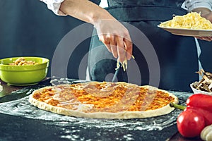 The chef in black apron makes pizza with his hands putting the ingredients for the pizza on the table.