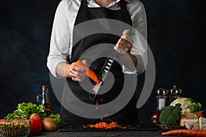 The chef in black apron shredding carrots with grater on black chopped board at professional restaurant kitchen on dark blue