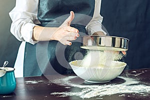 The chef in black apron sifts the flour through a sieve to prepare the dough for pizza