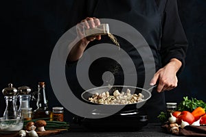 The chef in black uniform adds the pepper to the frying chicken in the wog pan isolated on the dark background. Restaurant`s dish