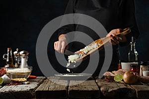 Chef in black uniform pours chopped onion from wooden board into plate on rustic table with ingredients background. Frozen motion