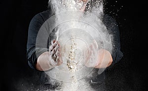 Chef in black uniform sprinkles white wheat flour in different directions, product scatters dust