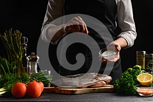Chef cooks fresh fish on the background with ingredients, salts it before cooking, freezing in motion, advertising photo for