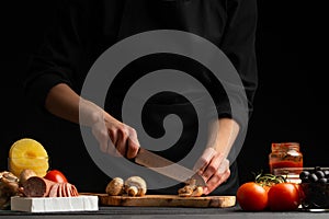 Chef cooks Italian pizza by slicing fresh mushrooms. Against the background of pizza ingredients. Black background, horizontal