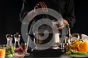 The chef cooks, sprinkling salt in a boiling pan, frozen in motion. Against the background of vegetables. Cooking and gastronomy,