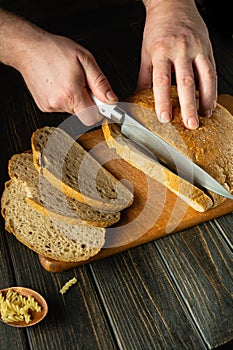 Chef cuts fresh bread with a knife on a kitchen board close-up. Slicing rye bread on the kitchen table or concept of healthy