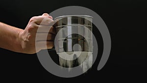 A chef hand holds a mug of sieve in hand and continuously sieving flour on a black background.