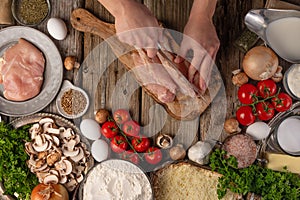 Chef hands cuts with knife chicken fillet for preparing meat filling on variety of ingredients background. Concept of cooking