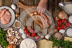 Chef hands cuts with knife chicken fillet on wooden chopped board with variety of ingredients background. Concept of cooking