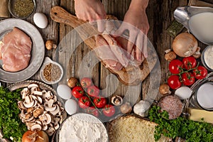 Chef hands cuts with knife chicken fillet on wooden table with variety of ingredients background. Concept of cooking process.