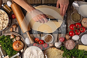 Chef hands cuts with knife the dough for pie on wooden table with variety of ingredients background. Concept of cooking process.