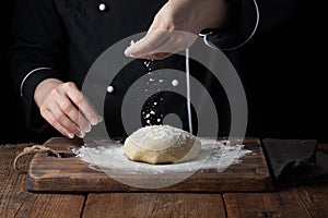 Chef hands pouring flour powder on raw dough using sieve on a black background, Cooking process