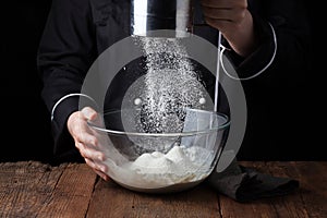 Chef hands pouring flour powder on raw dough using sieve on a black background, Cooking process