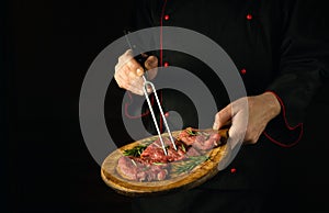 The chef holds a fork in his hand and cutting board with beef steaks before barbecue. Dark space for restaurant recipe or hotel
