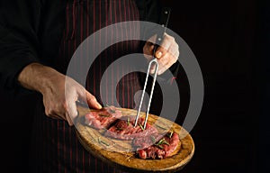 Chef holds a fork in his hand and a cutting board with beef steaks before barbecue. Dark space for restaurant recipe or hotel menu