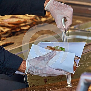 Chef with hygienic rubber gloves and a pair of tongs prepares a fish roll from a roll, a piece of mackerel fillet and a salad with