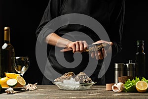 The chef opens the knife and cleans the raw oyster, on the background of white wine, lettuce, lemons and limes