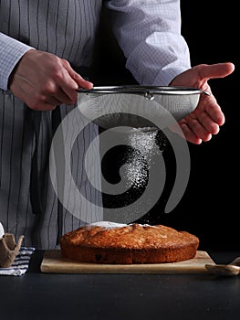 Chef pouring powdered sugar on tasty pie on dark background. ched processing pie or cake