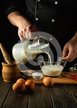A chef prepares a milkshake with fruits in the kitchen using a hand-held electric mixer. The cook pours milk into a deep plate.