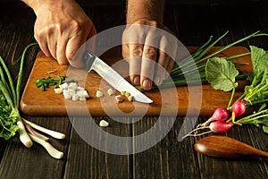 The chef prepares a salad in the kitchen of the restaurant. Close-up of the cook hands cutting green young onions with a knife on