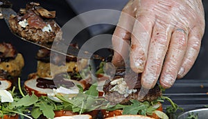 A chef preparing mini burgers for his customer, puts grilled meat on buns with vegetables.