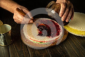 The chef is preparing a round waffle cake on the kitchen table. Adding jam with a spoon in the cook hand