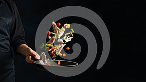 The chef preparing tasty dish with meat and mix of vegetables in a griddle, on a black background. Backstage of preparing