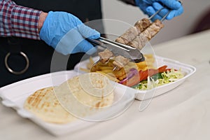 Chef put grilled meat into a lunch box with traditional Greek kalamaki dish. The cook takes pork fillet with tongs