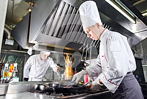 Chef in restaurant kitchen at stove with pan, cooking.