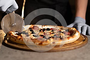 Chef& x27;s hand with a knife cuts hot pizza with olives and salami on the table on a wooden board in a pizzeria. Ready baked