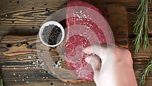 Chef salt a raw fresh steak with pepper and rosemary on wooden cutting board