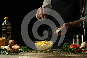 Chef salting french fries on a background with vegetables. Cooking tasty but harmful food