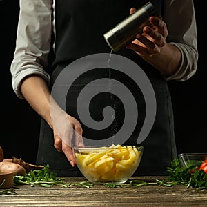 Chef salting french fries on a background with vegetables. Cooking tasty but harmful food