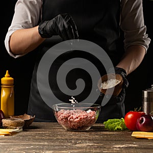 Chef salting minced meat to create a burger patty. Against the background with ingredients for a burger. Gastronomy, recipes,