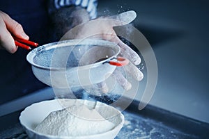 A chef sifts wheat flour through a large sieve into a white bowl, soiling his hands in flour. The process of preparing dough for